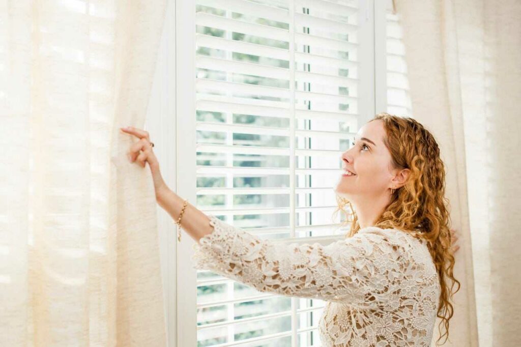 woman holding the curtain to see the custom blinds Blind Guy of Tri-Cities